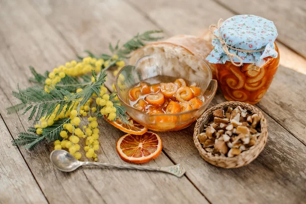 Composição da casca espiral de laranja cristalizada com xarope de açúcar em um frasco de vidro e prato perto do pires com nozes, mimosa, colher e pão — Fotografia de Stock