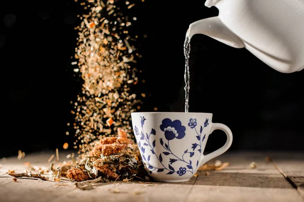 Чай наливается из белого чайника в стакан с голубым узором рядом с падающей горсткой сухих трав — стоковое фото