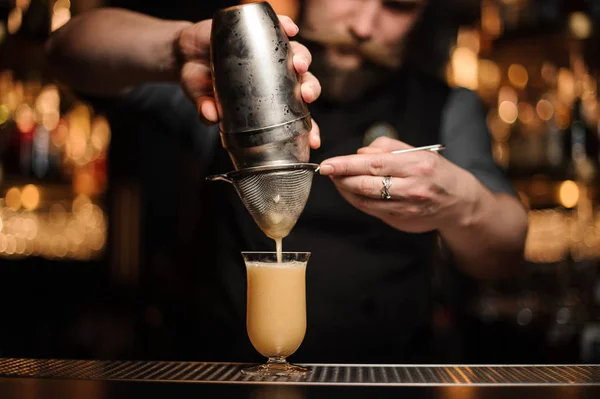 Barista con barba versando un delizioso cocktail dallo shaker in acciaio attraverso il setaccio — Foto Stock