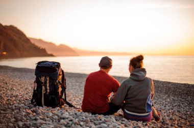 Sahilde oturan ve denize bakan bir sırt çantası ile erkek ve kadın