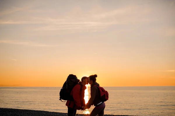 Par med rygg säckar stående nära havet i solnedgången håller händer och kyssar — Stockfoto