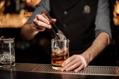 Profesyonel barmen kahverengi bir kokteyl ile ölçüm cam fincan büyük bir buz küpü ekleyerek