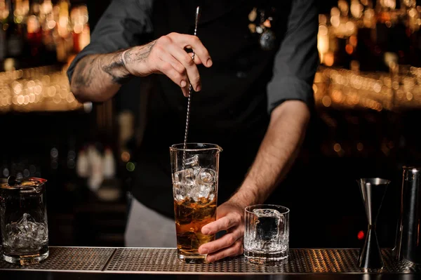 Barman brassant de nombreux glaçons dans le grand verre avec un cocktail brun avec une cuillère — Photo