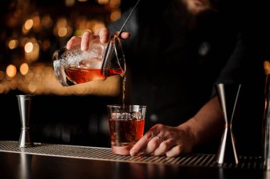 Barmen bir büyük buz küpü ile cam süzgeci aracılığıyla ölçüm bardaktan kırmızı alkol dökme