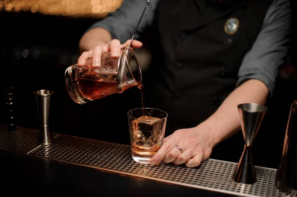 Cantinero vertiendo un alcohol de la taza de medir a través del colador al vaso con un cubo de hielo grande — Foto de Stock