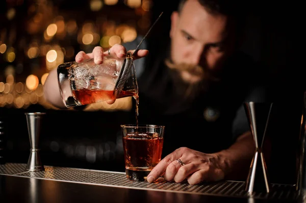 Cantinero profesional que vierte un alcohol de la taza de medir a través del colador al vaso con un cubo de hielo grande — Foto de Stock