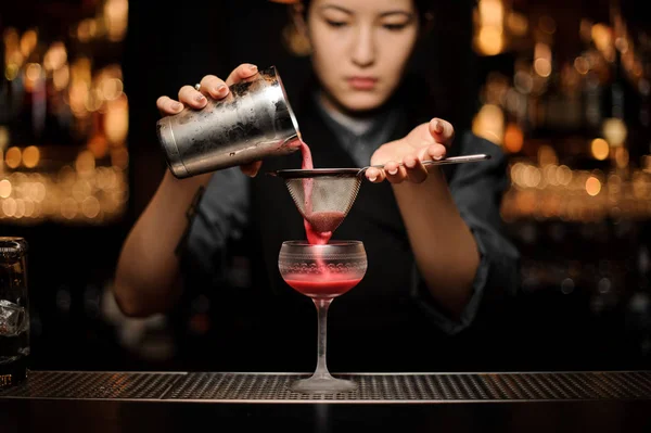 Professional bartender girl pouring a smooth crimson cocktail through the sieve to the glass — Stock Photo, Image