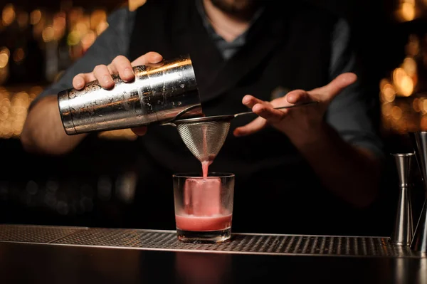 Barman derramando um coquetel rosa suave através da peneira para o vidro com um grande cubo de gelo — Fotografia de Stock