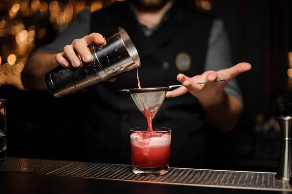 Cantinero profesional vertiendo un cóctel rosa suave a través del tamiz al vaso con un cubo de hielo grande — Foto de Stock