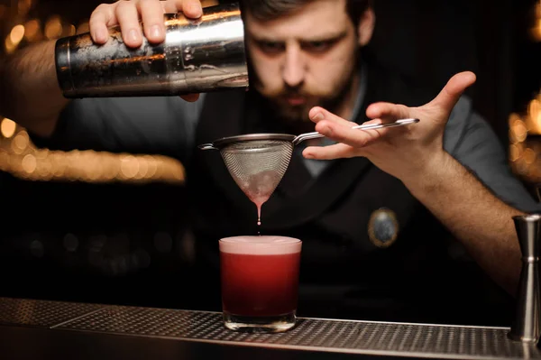 Barman avec une barbe versant un cocktail rose lisse à travers le tamis au verre avec un gros glaçon — Photo