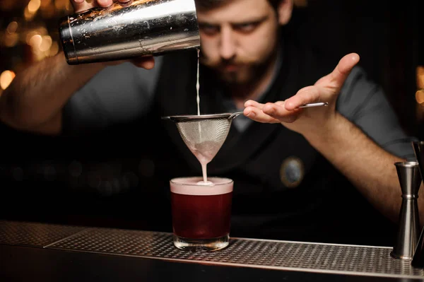 Barman com uma barba derramando um coquetel carmesim suave através da peneira para o vidro com um grande cubo de gelo — Fotografia de Stock