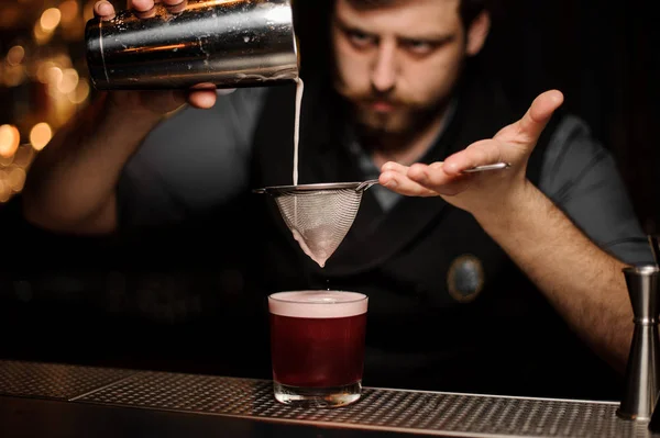 stock image Bartender with a beard pouring a smooth dark red cocktail through the sieve to the glass with one big ice cube