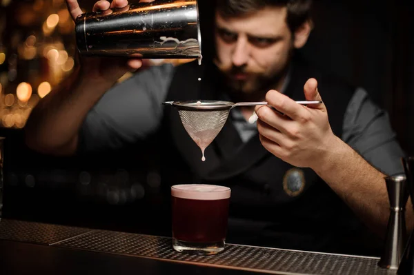 Barista con la barba versando un cocktail rosa liscio attraverso il setaccio al bicchiere dallo shaker — Foto Stock