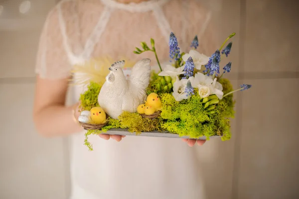 Menina segurando uma composição de primavera com chiken de brinquedo e flores brancas e azuis — Fotografia de Stock