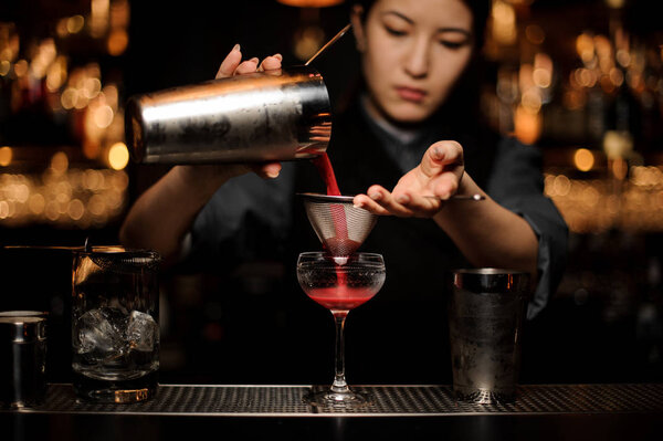 Female bartender pouring cocktail with shaker and sieve