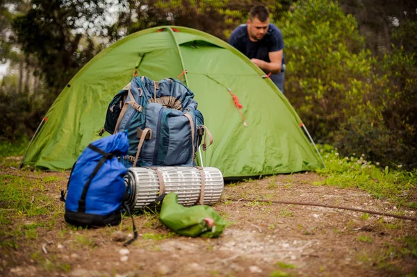 Young man sets up a green tent near the hiking equipment