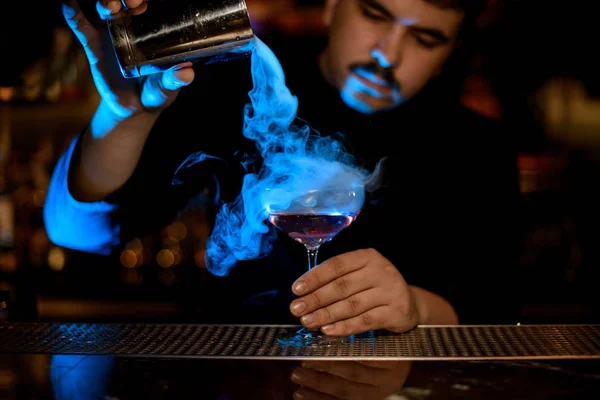 Professionelle männliche Barkeeper gießt einen Rauch in das Cocktailglas aus dem Stahlshaker unter dem blauen Licht — Stockfoto