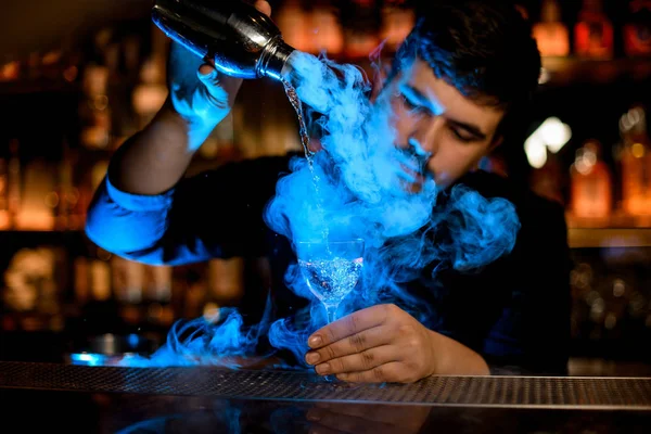 Professionelle männliche Barkeeper gießt einen Rauch in das Cocktailglas aus dem Shaker unter dem blauen Licht — Stockfoto