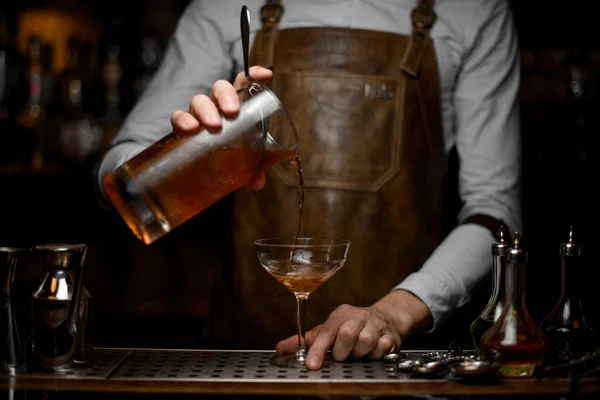 Professioneller männlicher Barkeeper, der ein braunes alkoholisches Getränk aus dem Messbecher in das Cocktailglas gießt — Stockfoto