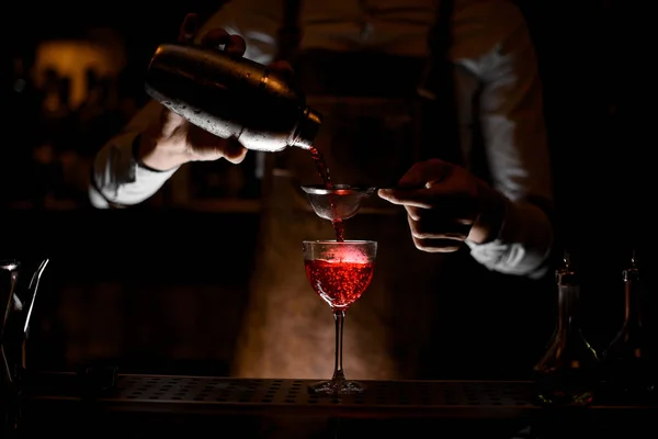 革のエプロンのバーテンダーは、暗闇の中でふるいを通してスチールシェーカーから赤いアルコール飲料を注ぎます — ストック写真