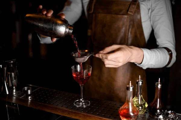 Barman in lederen schort het gieten van een rode alcoholische drank uit de stalen Shaker door de zeef — Stockfoto