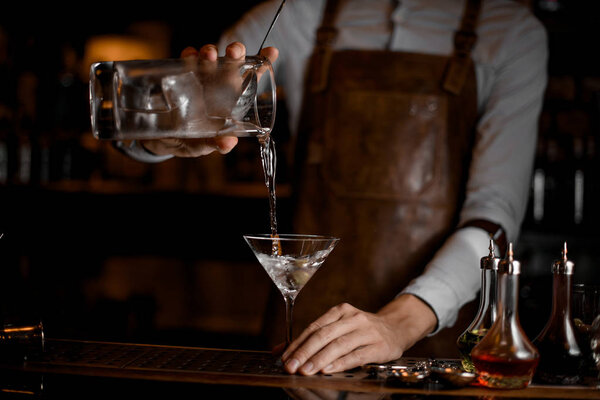 Профессиональный бармен наливает прозрачный алкогольный напиток из мерного стакана в бокал мартини
