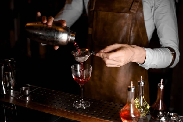 Barman profissional derramando uma bebida alcoólica vermelha da coqueteleira de aço através da peneira — Fotografia de Stock