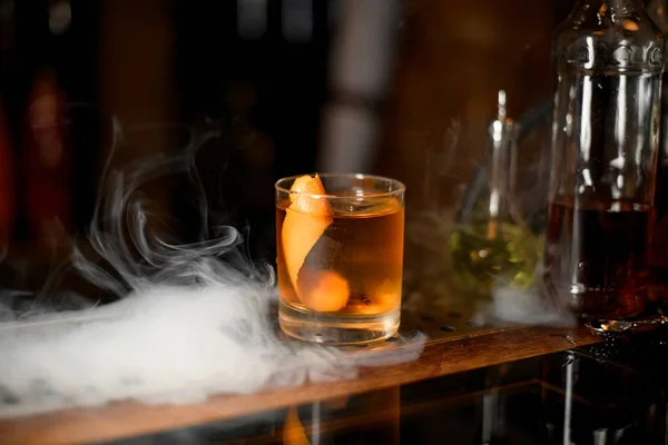 Golden cocktail in the glass with one ice cube and orange zest in the smoke