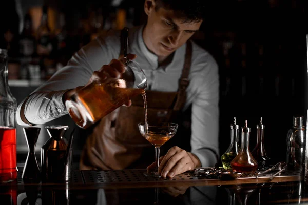 Barman gieten een bruine alcoholische cocktail van de maatbeker naar het glas in het donker — Stockfoto