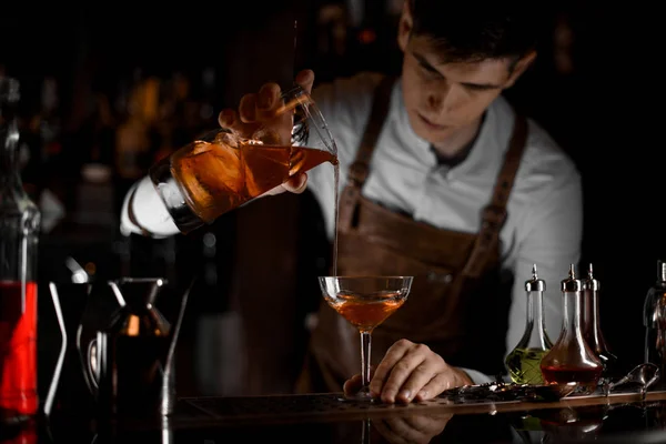 Attraktiver Barkeeper, der im Dunkeln einen braunen alkoholischen Cocktail aus dem Messbecher ins Glas gießt — Stockfoto