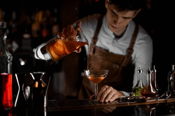 Barman masculino atraente derramando um coquetel alcoólico marrom do copo de medição para o vidro no escuro — Fotografia de Stock