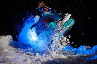 Turuncu ve mavi spor giyim giymiş Snowboarder kız kar üzerinde hileler yapar