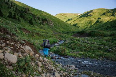 Yürüyüş sırt çantası ve dere boyunca sopalarla dağ nehri yakınında yürüyen adam
