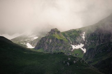 Beyaz sisin altındaki kayalık dağların manzarası kar kalıntılarıyla kaplı çimlerle kaplı