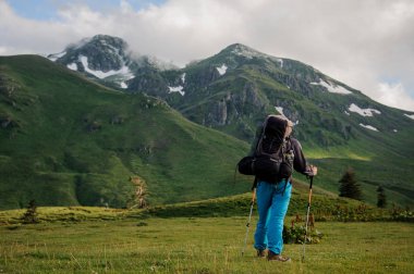 Tepede duran arka görüşteki adam, kar kalıntılarıyla kaplı çimenlerle kaplı kayalık dağlara bakıyor.