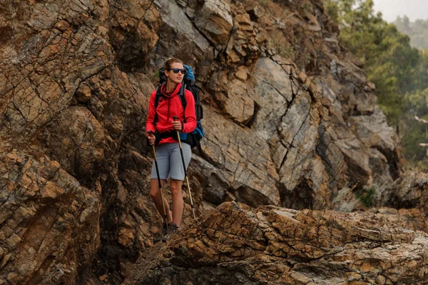 Glimlachend meisje staande op de rotsen met wandelrugzak en wandelstokken — Stockfoto