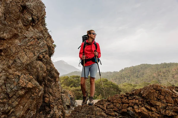 Donna in occhiali da sole in piedi sulla roccia con zaino da trekking e bastoni da passeggio — Foto Stock