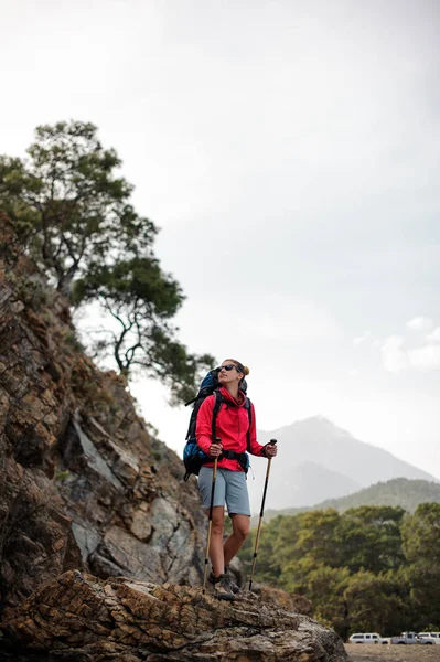 Slim fit woman in sportwear standing with hiking backpack and sticks on the rock and looking up