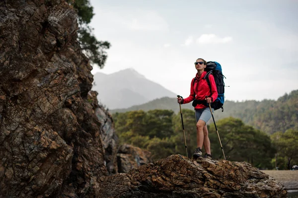 Donna slim fit in abbigliamento sportivo e occhiali da sole in piedi con zaino da trekking e bastoni sulla roccia — Foto Stock