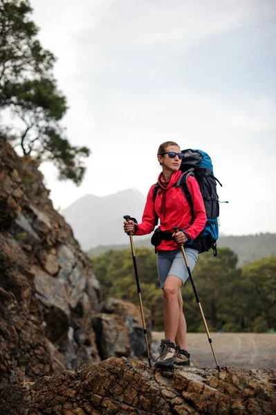 Mujer en ropa especial de pie con mochila de senderismo y palos en la roca —  Fotos de Stock