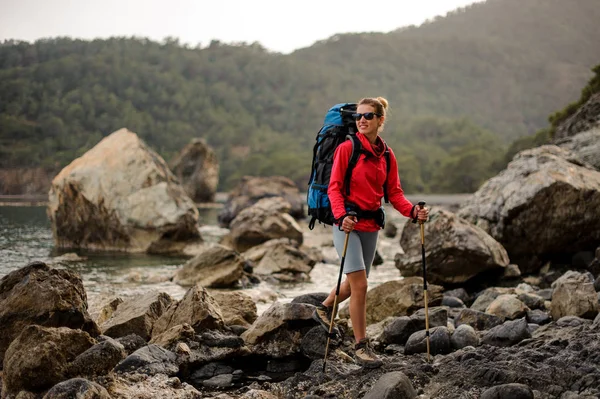 Chica de pie sobre las rocas en la costa en el mar con mochila de senderismo —  Fotos de Stock