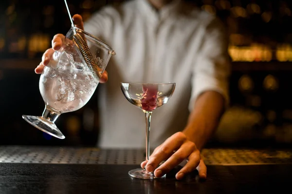 Barman masculino derramando um coquetel do copo de medição com um filtro para um copo com cubo de gelo rosa — Fotografia de Stock