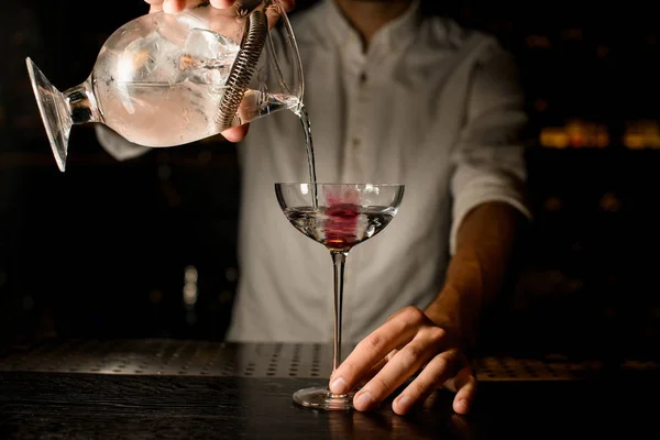 Профессиональный бармен наливает коктейль из мерной чашки с фильтром в стакан с розовым кубиком льда — стоковое фото