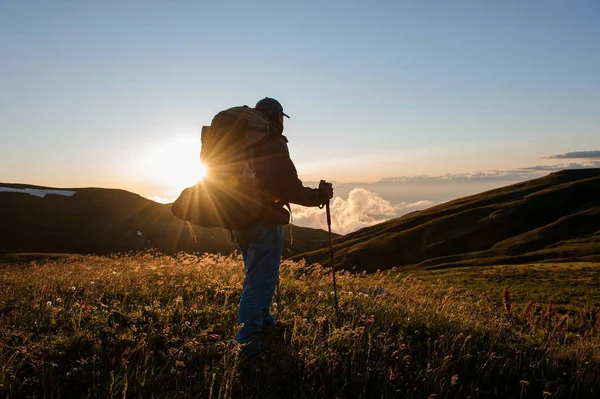 Hombre vista trasera de pie en el campo de la colina con mochila de senderismo y palos mirando al sol —  Fotos de Stock