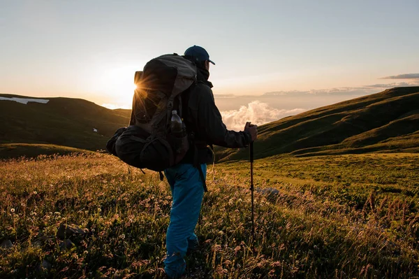 Hombre vista trasera de pie en el campo de la colina con mochila de senderismo y palos mirando al amanecer —  Fotos de Stock