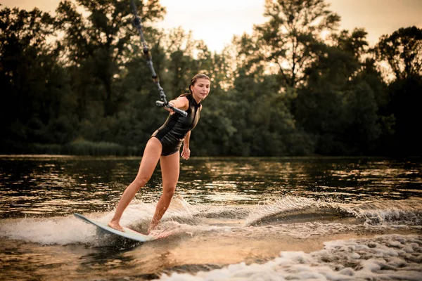 Girl riding on the wakeboard on the river in the sunset holding a rope of the motorboat — Stock Photo, Image