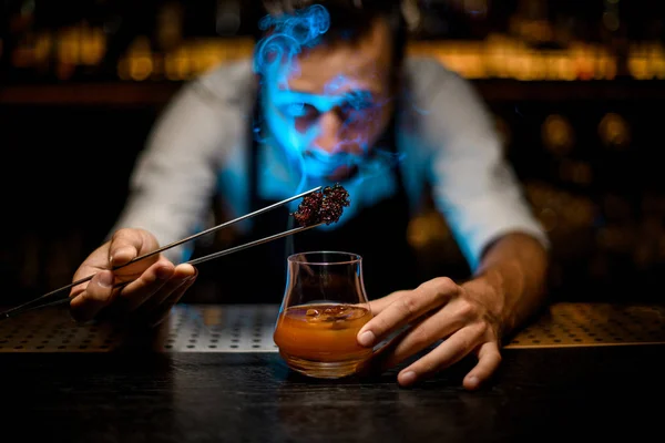 Cantinero masculino añadiendo caramelo fundido refrigerado con pinzas al cóctel con cubitos de hielo bajo luz azul — Foto de Stock