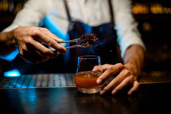 Professioneller Barkeeper, der dem Cocktailglas mit Eiswürfeln unter blauem Licht gekühltes, schmelzendes Karamell mit Twezzers hinzufügt — Stockfoto
