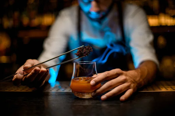 Barman masculino profesional añadiendo caramelo fundido refrigerado con pinzas a la copa de cóctel con cubitos de hielo bajo luces azules — Foto de Stock