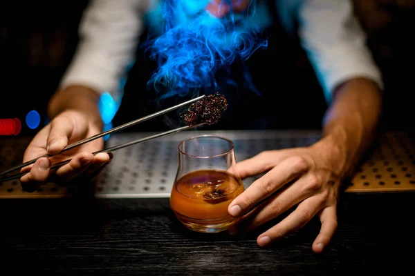 Bartender adding chilled melting caramel with twezzers to the cocktail with ice cubes under blue lights — Stock Photo, Image